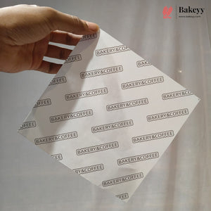 A hand holds a square white bakery gift wrap or napkin printed with a repeating 'BAKERY&COFFEE' logo pattern, set against a soft, light gray draped fabric background.