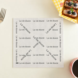 A square white placemat printed with the phrase 'La vie douce' repeated in black at the borders and diagonally across the center, styled on a light beige tabletop with a fork, blueberry toast, and coffee cup nearby.