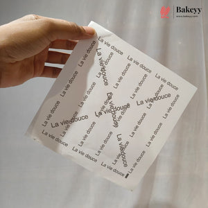 A hand holds a white sheet of paper printed with the repeating phrase 'La vie douce' in black script, photographed against a light gray background. A Bakeyy logo appears in the top right corner.