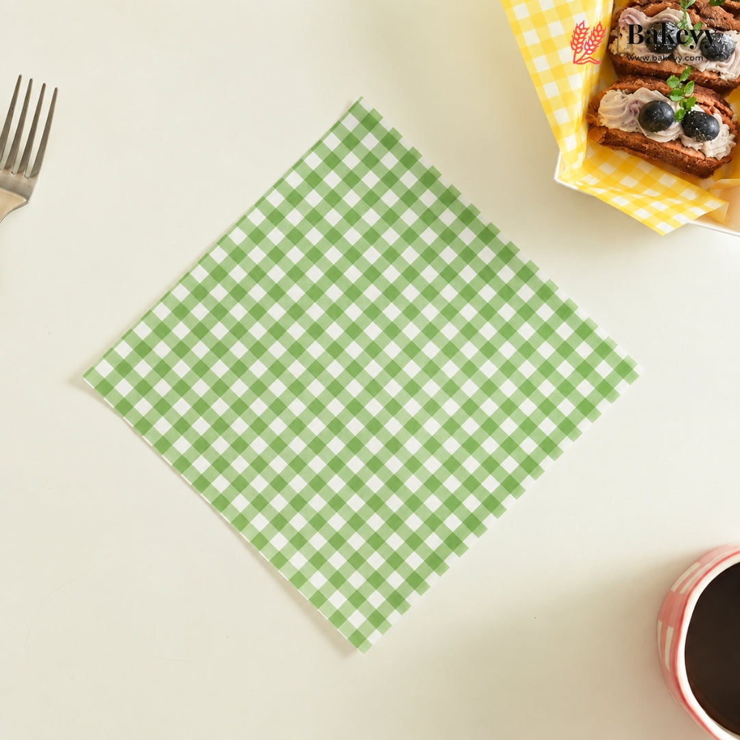 A green and white gingham checkered paper napkin is laid flat on a light cream tabletop. A fork is partially visible on the left, and a small basket of pastries and a cup are cropped on the right and bottom edges, creating a styled flat-lay scene.