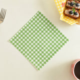 A green and white gingham checkered paper napkin is laid flat on a light cream tabletop. A fork is partially visible on the left, and a small basket of pastries and a cup are cropped on the right and bottom edges, creating a styled flat-lay scene.