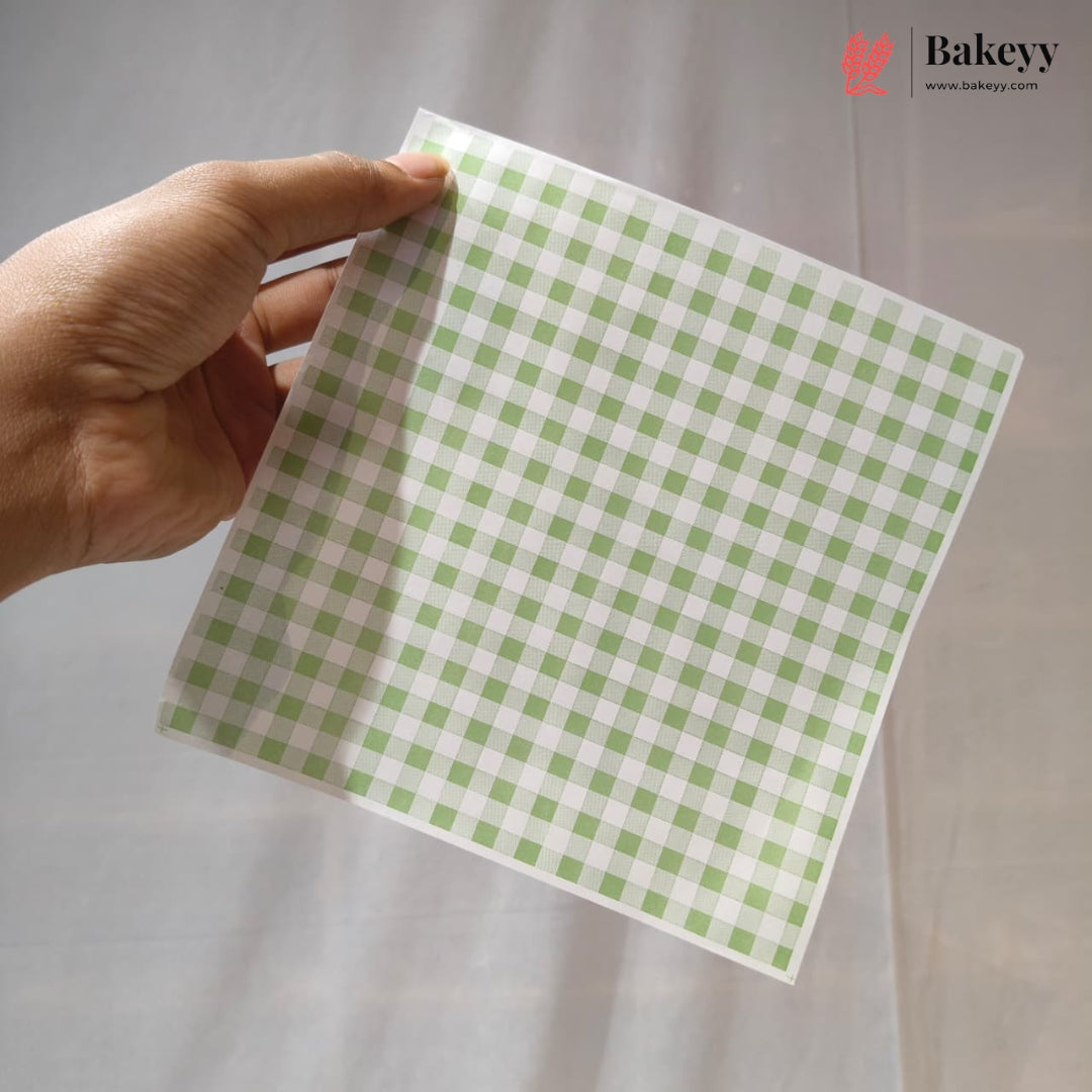 A hand holding a square sheet of white washi tape with a light green gingham checkered pattern, set against a soft white fabric background. The Bakeyy logo is visible in the top right corner.