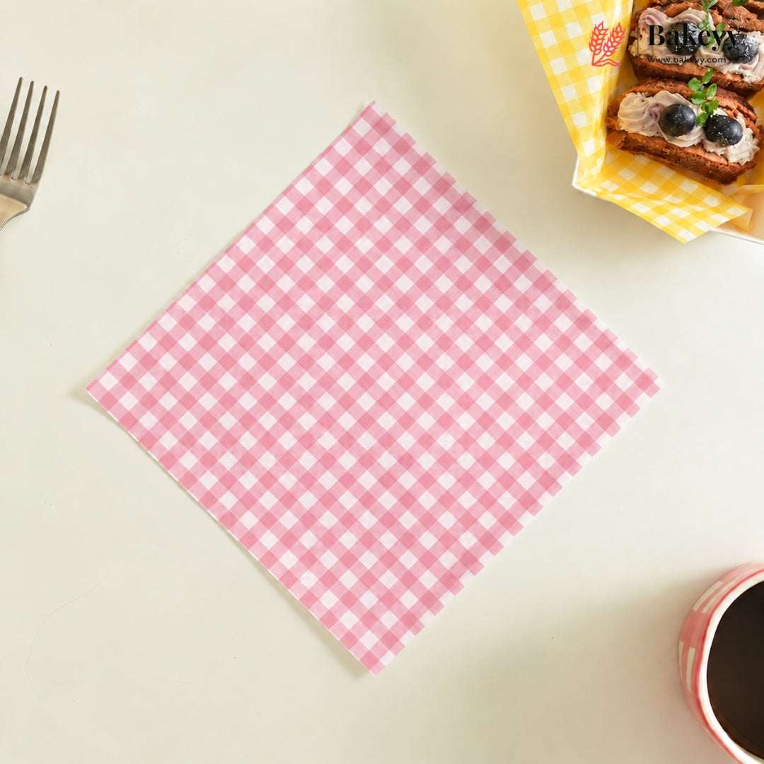A pink and white gingham checkered square napkin is laid flat on a light beige tabletop, with a fork at the top left, a basket of pastries at the top right, and a cup at the bottom right.