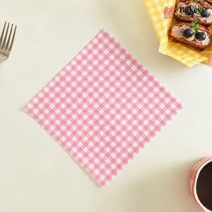 A pink and white gingham checkered square napkin is laid flat on a light beige tabletop, with a fork at the top left, a basket of pastries at the top right, and a cup at the bottom right.