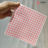 A hand holds a square sheet of pink and white gingham checkered paper against a light gray fabric background. A Bakeyy logo is visible in the top right corner.