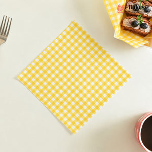 A square yellow and white gingham checkered napkin is displayed diagonally on a light cream-colored tabletop. A fork is visible on the left, and a basket of pastries with a 'BakeY' logo on the napkin is in the top right corner.