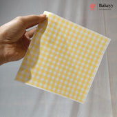 A hand holding a square sheet of yellow and white gingham checkered baking paper against a light gray fabric backdrop. The Bakeyy logo is visible in the top right corner.