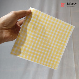 A hand holding a square sheet of yellow and white gingham checkered baking paper against a light gray fabric backdrop. The Bakeyy logo is visible in the top right corner.