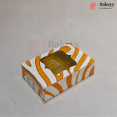 A small rectangular bakery box with a zebra-like orange and white pattern, featuring a clear plastic window on top that reveals a golden-brown tray of pastries inside. The box is photographed on a plain light gray background with faint Bakeyy watermarks.