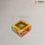A square decorative bakery box with a clear plastic window on top, showing a gold-colored tray inside. The box features a colorful abstract pattern in orange, yellow, red, and green on a light gray background with Bakeey watermarks.