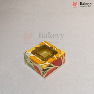 A square decorative bakery box with a clear plastic window on top, showing a gold-colored tray inside. The box features a colorful abstract pattern in orange, yellow, red, and green on a light gray background with Bakeey watermarks.
