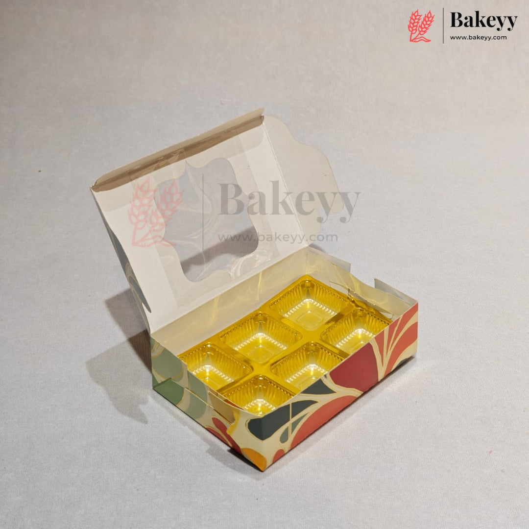 An open bakery box with a hinged lid, featuring a gold foil crumb tray inside and a printed multicolor pattern on the exterior. The Bakeyy logo is visible in the top right corner.