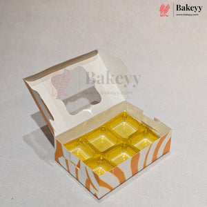 An open cardboard bakery box with a hinged lid and six clear plastic cup compartments lined with gold foil, shown on a light gray background. The box has orange and white wavy stripes on the lower side panel, and a watermark for Bakeyy is visible.