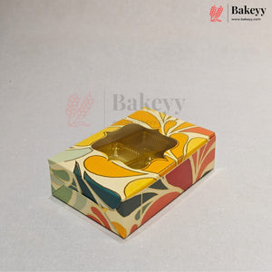 A rectangular bakery box with a colorful retro floral pattern in yellow, orange, red, green, and beige, featuring a clear window showing golden cupcakes inside. The image has a Bakeyy watermark and logo in the top right corner.