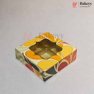 A square, multi-compartment gift box with a colorful retro floral pattern in yellow, orange, red, and green on a cream background, featuring a clear window on top. The Bakeyy brand logo is visible in the top right corner.