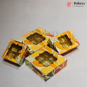 Four decorative packaging boxes with a retro floral pattern in yellow, orange, green, and red, each featuring a clear window showing gold-colored cupcake liners inside, set against a plain light gray background.