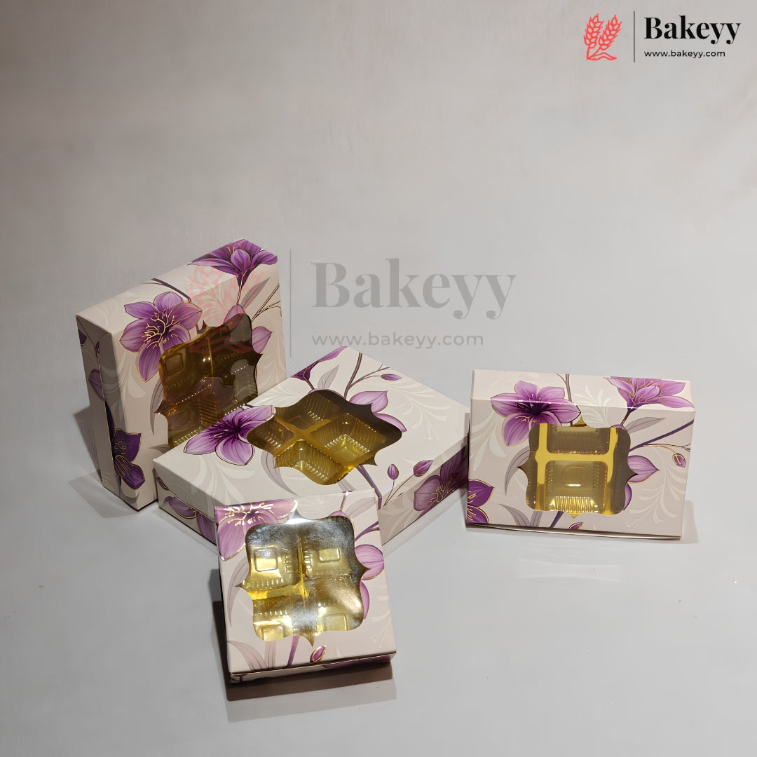 Four decorative rectangular boxes with purple floral prints and clear windows showing gold foil-wrapped items inside, arranged on a light gray surface. A Bakeyy watermark/logo appears in the top right and across the center.