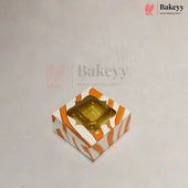 A square bakery box with a clear plastic window on top, decorated with orange and white diagonal stripes, shown on a light gray background with Bakeyy branding.