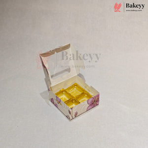 A small open bakery box with a clear window and a yellow tray inside, decorated with a purple floral pattern on a light beige background. The Bakeyy logo is visible in the top right corner.