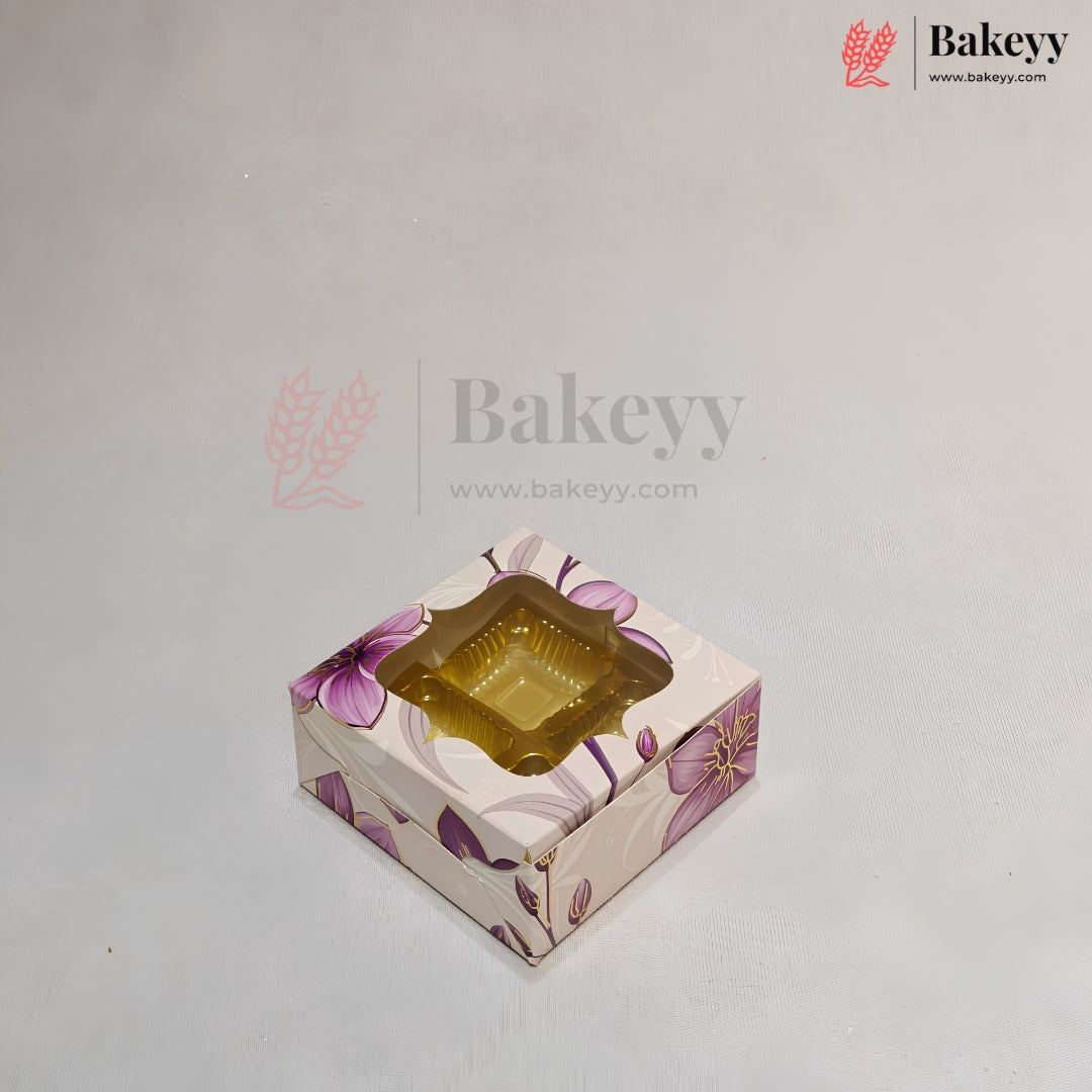 A square bakery box with a floral pattern in purple and beige, featuring a clear plastic window on top displaying a golden-brown pastry, set against a plain light gray background.