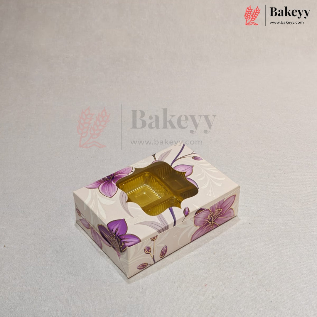 A rectangular bakery box with a floral pattern in purple and pink on a cream background, featuring a clear plastic window on top showing a golden-brown pastry inside. The image has a watermark for 'Bakeyy' on a light gray background.