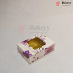 A rectangular bakery box with a floral pattern in purple and pink on a cream background, featuring a clear plastic window on top showing a golden-brown pastry inside. The image has a watermark for 'Bakeyy' on a light gray background.