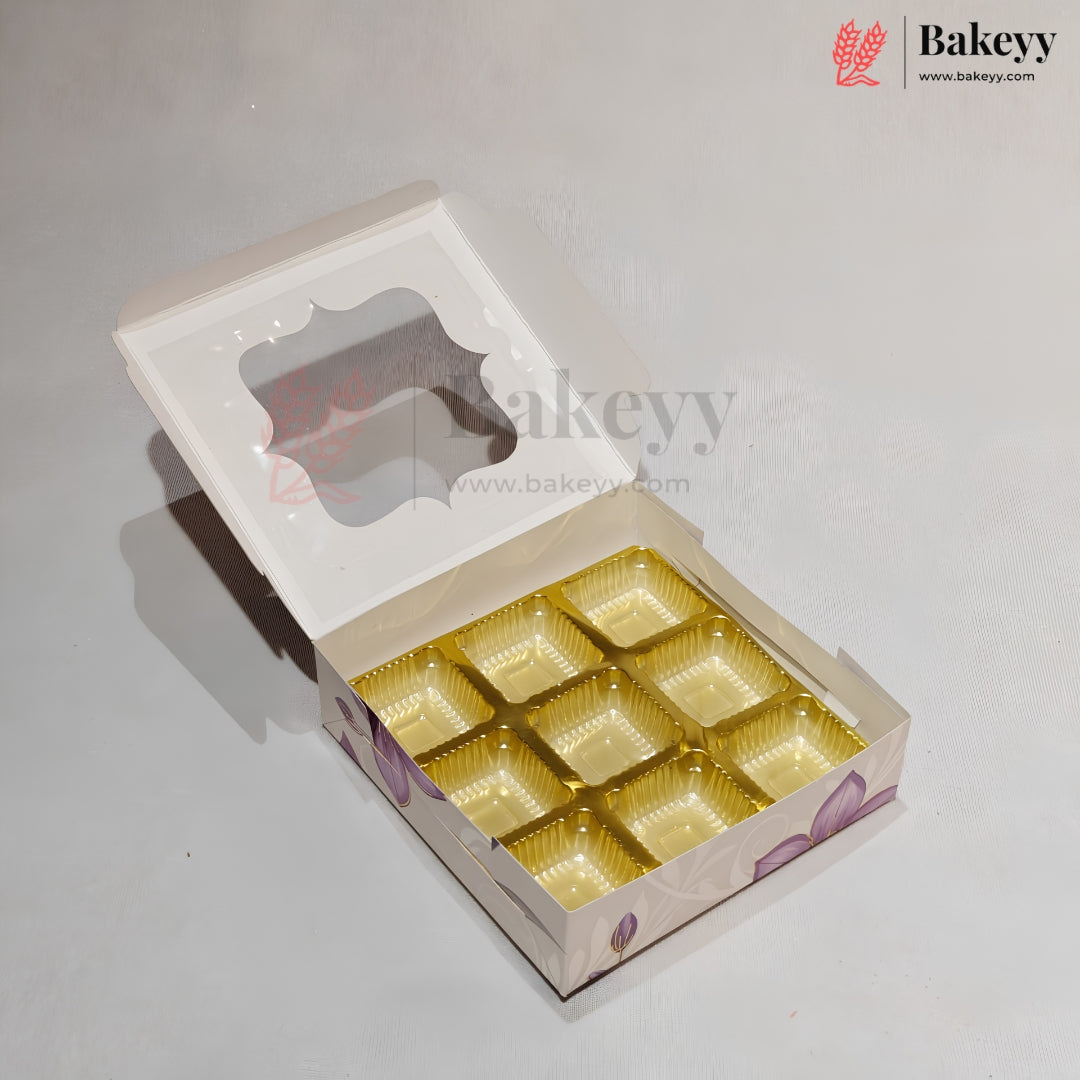 An open white bakery box with a clear window on the lid and a gold foil tray insert holding multiple square compartments, photographed on a light gray background. A Bakeyy logo watermark is visible in the top right and across the center.