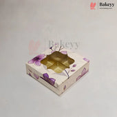 A square bakery gift box with a floral pattern in purple and beige, featuring a clear window that reveals a gold-colored tray inside, set against a light gray background.