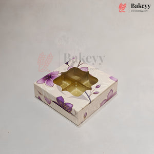 A square bakery gift box with a floral pattern in purple and beige, featuring a clear window that reveals a gold-colored tray inside, set against a light gray background.