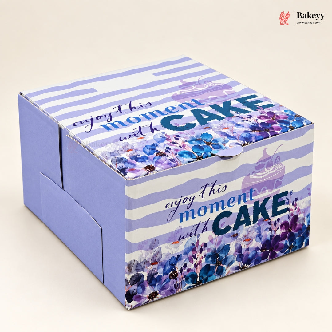 A square cardboard gift box with a purple and white striped pattern, decorated with blue and purple floral designs and the text 'enjoy this moment with CAKE'. The box is photographed on a light beige background with a Bakeyy logo in the top right corner.