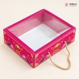 A decorative pink and gold rectangular gift box with a clear plastic window on top, shown on a light beige background. The box features festive illustrations, a gold rope handle, and a Bakeyy logo watermark in the top right corner.