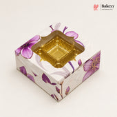 A square bakery box with a floral purple and white pattern, featuring a clear window that reveals a gold-colored square tray inside, set against a plain off-white background.