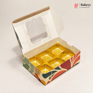 An open bakery box with a clear window on the lid and six gold foil compartments inside, shown on a light beige background. The box has a colorful abstract pattern in green, red, orange, and beige.