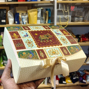 A hand holds a decorated rectangular gift box with a lid featuring a colorful patchwork design of an elephant and peacocks, tied with a cream ribbon, against a blurred bakery shelf background.