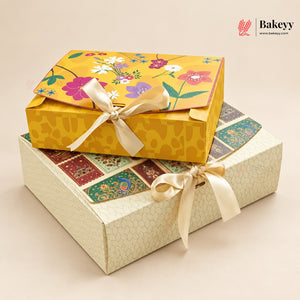Two decorative gift boxes stacked on a light beige background. The top box is mustard yellow with a floral print and tied with a cream ribbon, while the bottom box is cream-colored with a subtle honeycomb pattern and patchwork-style panels on the lid, also tied with a cream ribbon.