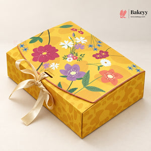A rectangular yellow bakery gift box with a floral-patterned lid, tied with a cream satin ribbon, photographed on a light beige background. The Bakeey logo appears in the top right corner.