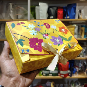 A hand holds a yellow gift box decorated with a colorful floral pattern and tied with a cream ribbon, set against a blurred background of shelves with bags.