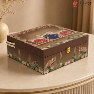 A rectangular decorative jewelry box with a dark brown base and intricate multicolor printed illustrations of peacocks, elephants, flowers, and traditional motifs on the lid and sides. The box has gold-tone metal latches and is displayed on a beige marble table with a warm, elegant background.