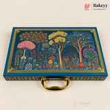 A rectangular blue decorative gift box with a gold handle, featuring an illustrated forest scene with colorful trees and floral motifs on the lid, photographed on a light beige background. The Bakeyy logo appears in the top right corner.