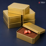 Six square gold metal cake or chocolate boxes are arranged on a dark blue background, with one box open in front showing cocoa powder and strawberry slices inside. A Bakeyy logo appears in the top right corner.