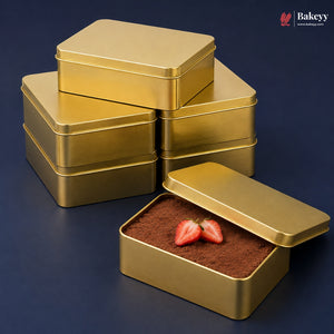 Six square gold metal cake or chocolate boxes are arranged on a dark blue background, with one box open in front showing cocoa powder and strawberry slices inside. A Bakeyy logo appears in the top right corner.
