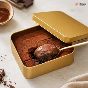 A square gold metal tin filled with chocolate dessert, with the lid partly open and a spoon lifting a scoop of the chocolate cake. The scene is styled on a light marble surface with cocoa powder, chocolate pieces, and a small bowl in the background.