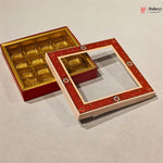 A red and gold square chocolate box with an open lid, shown on a light gray background. The box has a red patterned outer frame with a clear window and gold-colored compartments inside.
