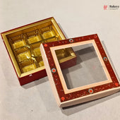 A decorative red and gold square gift box shown open, with a gold foil tray inside and the lid leaning against it. The lid has a clear window surrounded by ornate red floral detailing and a pale pink border.