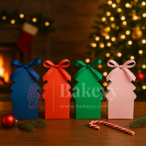 Four colorful Christmas tree-shaped boxes with ribbons on a wooden table, decorated with a Christmas tree and candy cane in the background.