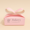 Pink gift box with a ribbon on a beige background, featuring the brand 'Bakeyy'.