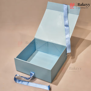 A light blue square gift box with its lid propped open, featuring a matching satin ribbon tied around the exterior. The box is photographed on a warm beige background with a visible Bakeyy watermark.