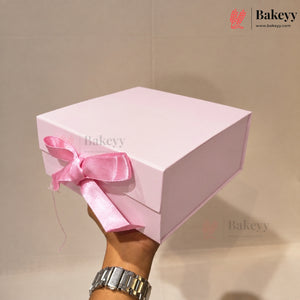 A hand holding a light pink square gift box tied with a matching pink satin ribbon bow, against a plain beige background. The Bakeyy logo is visible in the top right corner.