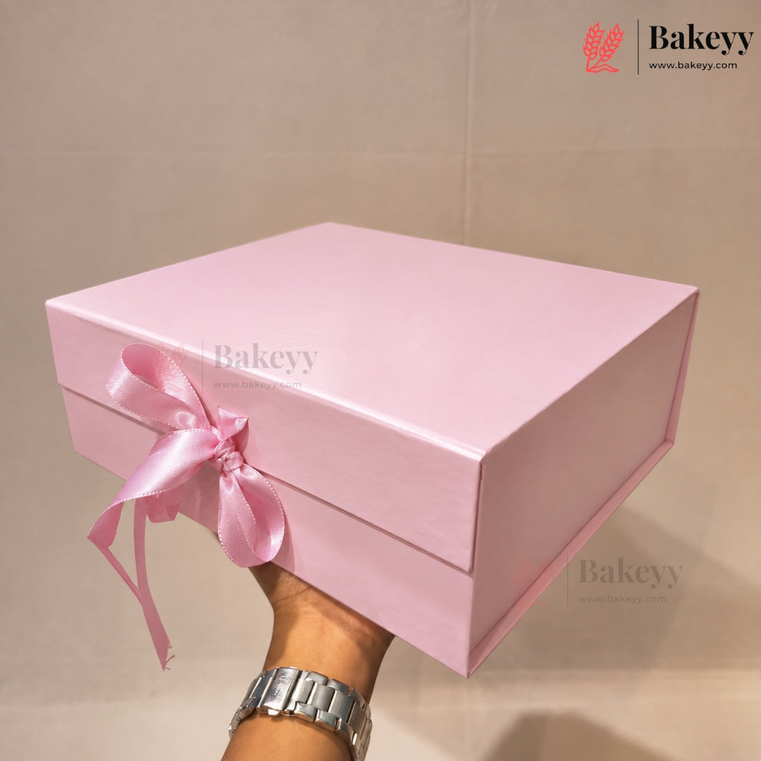 A light pink rectangular gift box with a matching pink satin ribbon bow, held by a hand wearing a silver watch against a beige background. The Bakeyy logo is visible in the top right corner.