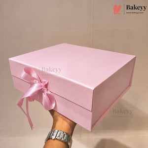 A light pink rectangular gift box with a matching pink satin ribbon bow, held by a hand wearing a silver watch against a beige background. The Bakeyy logo is visible in the top right corner.